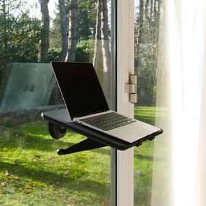 May include: A black laptop stand with a laptop on it, mounted to a window frame. The stand has a black base and a black arm that extends out to hold the laptop. The laptop is open and has a black screen. The window is open and there is a view of green grass and trees outside.