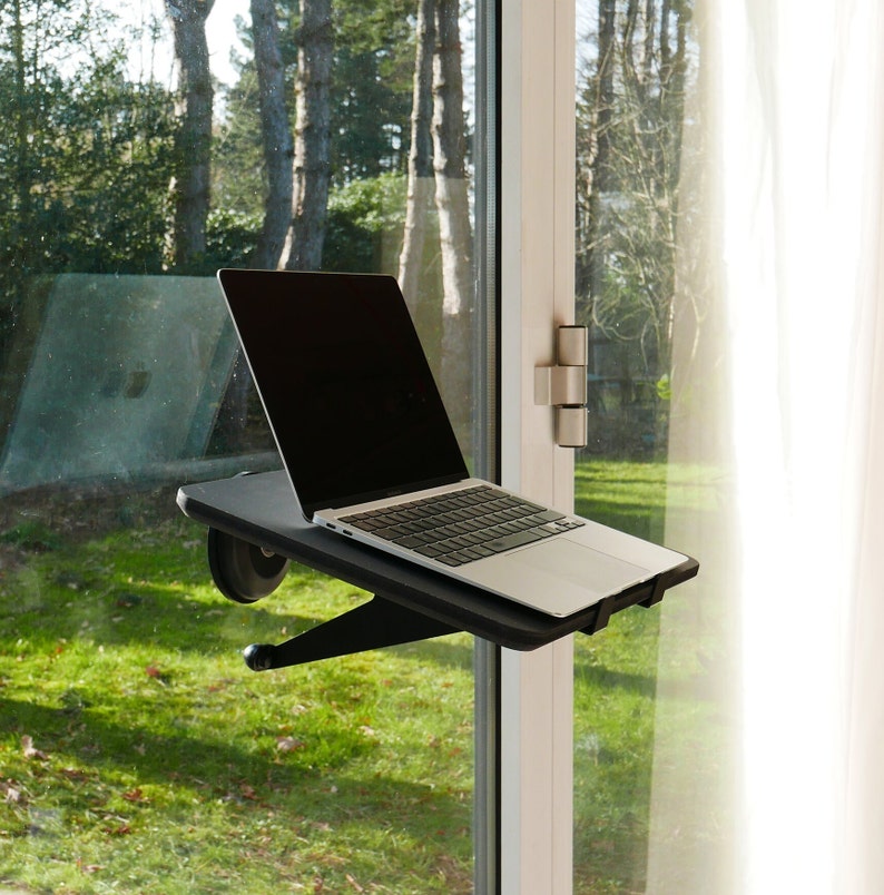 May include: A black laptop stand with a laptop on it, mounted to a window frame. The stand has a black base and a black arm that extends out to hold the laptop. The laptop is open and has a black screen. The window is open and there is a view of green grass and trees outside.
