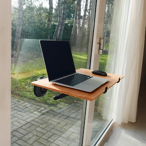 May include: A laptop computer sits on a wooden desk that is attached to a window. The desk is made of light brown wood and has a black metal frame. The laptop is open and has a black screen. A wireless mouse is on the desk.