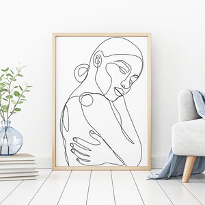 May include: Line art drawing of a person's face and shoulders in a minimalist style. The drawing is in black and white and is framed in a light wood frame.