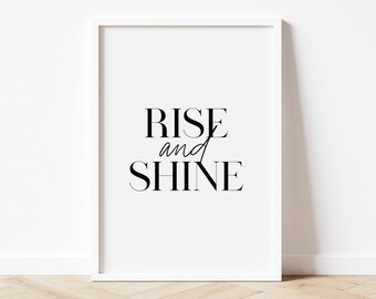 May include: A white framed print with the text "Rise and Shine" in black lettering. The print is on a white background and is sitting on a light wood floor.