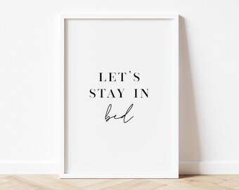 May include: White framed print with black text that reads "Let's Stay In Bed" on a white background. The print is on a wooden floor.
