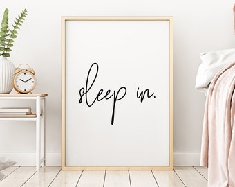 May include: A gold framed print with the text "sleep in." hanging on a white wall in a bedroom setting. The print is above a white nightstand with a gold alarm clock and a vase with green plants.