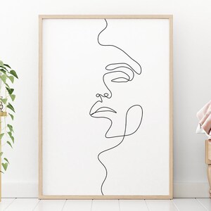 May include: A black and white line drawing of a face in a simple, minimalist style. The drawing is framed in a light wood frame and is hanging on a white wall.