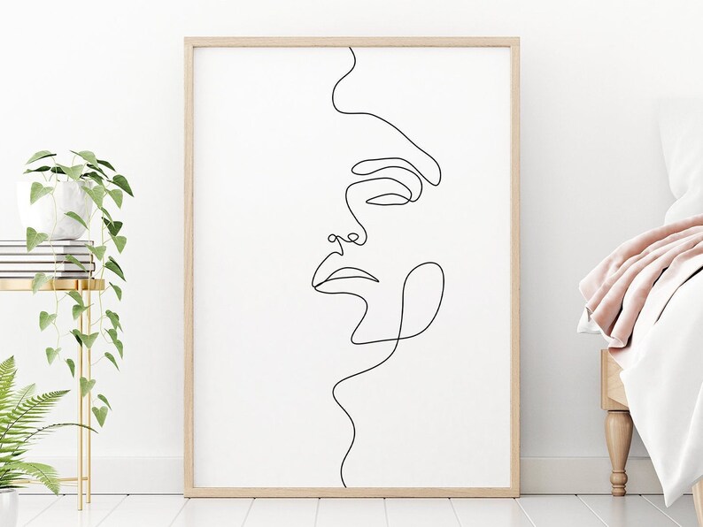 May include: A black and white line drawing of a face in a simple, minimalist style. The drawing is framed in a light wood frame and is hanging on a white wall.