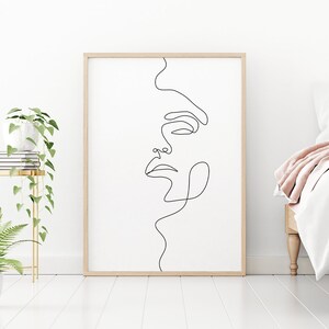 May include: A black and white line drawing of a face in a simple wooden frame. The artwork is hanging on a white wall above a white bed with a pink throw blanket.