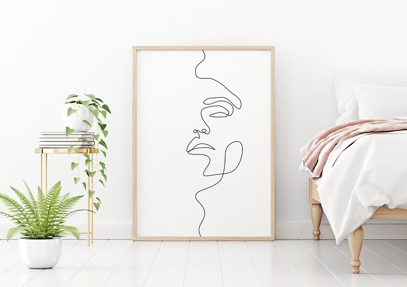May include: A black and white line drawing of a face in a simple wooden frame. The artwork is hanging on a white wall above a white bed with a pink throw blanket.