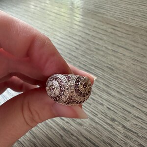 May include: A silver-toned ring with a rounded, textured design. The ring is encrusted with small, round, clear and dark purple stones, creating a spiral pattern. The ring is held by a hand.