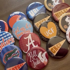 May include: A collection of circular pin-back buttons featuring various collegiate sports themes, including pennant designs, team logos, and text such as Florida, URI, UCF, Roll Tide, and Get up it's gameday, displayed in a variety of colors and patterns.