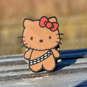 May include: A brown and black Hello Kitty character figurine with a red bow. The character is dressed as Chewbacca from Star Wars, with a brown fur coat and a white bandolier.