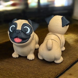 Puede incluir: Dos figuritas de pug impresas en 3D, una mirando hacia adelante y la otra hacia atrás. Ambas son de color beige con orejas y narices negras, y ojos blancos. Las figuritas están sentadas sobre una alfombra marrón.