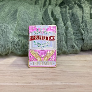 May include: Pink and gold enamel pin with a tarot card design. The pin features the word "SMUT" in a red banner, with "THE READER" at the bottom. The design includes open hands, leaves, and hearts.
