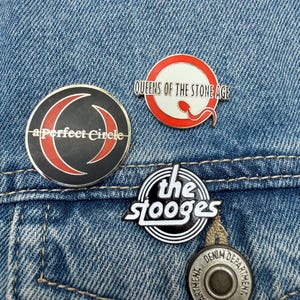 May include: Three enamel pins with band logos on a denim jacket. The pins display the logos of "a perfect Circle", "Queens of the Stone Age", and "the stooges". The pins have red, black, and white designs, and are attached to a blue denim jacket.