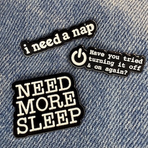 May include: Three black and white enamel pins with humorous phrases. One pin reads "i need a nap", another says "Have you tried turning it off & on again?", and the third says "NEED MORE SLEEP". The pins are on a denim background.