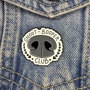 May include: A circular enamel pin, black and white, with the words "SNOOT-BOOPER CLUB" around a grey dog nose design. The pin is attached to a light blue denim jacket.