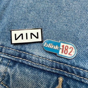 May include: Two enamel pins on a denim background. One rectangular pin features the letters "NIN" in black on white. The other, pill-shaped pin has "blink-182" and "182" in red and white on a blue background.