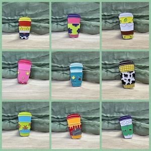 May include: Nine enamel pins in the shape of coffee cups, each with a different design. The pins feature various patterns, including flowers, stars, and a cow print. The background is a green fabric, and the pins are brightly coloured.