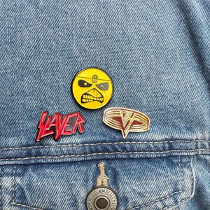 May include: Three enamel pins on a denim jacket. One pin is a yellow circle with a black and yellow face. The second pin is red text that says "SLAYER". The third pin is a gold and red design.