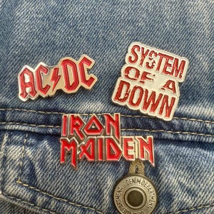 May include: Three enamel pins with red and silver detailing are displayed on a denim jacket. The pins feature the logos of the bands AC/DC, System of a Down, and Iron Maiden. The jacket has a button that says "Denim Department."