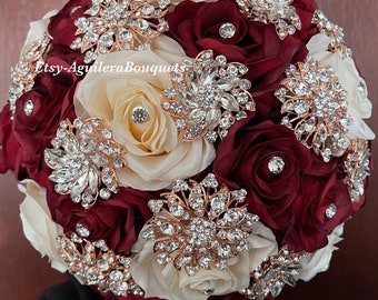May include: A bridal bouquet made with ivory and burgundy silk roses, adorned with gold and rhinestone brooches. The bouquet is a beautiful and elegant choice for a wedding.