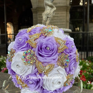 Pu&ograve; includere: Un bouquet da sposa fatto di rose bianche e lavanda con spille a forma di farfalla dorate e strass. Il bouquet &egrave; su un piatto di vetro trasparente. Il testo "Etsy-AguileraBouquets" &egrave; sul bouquet.