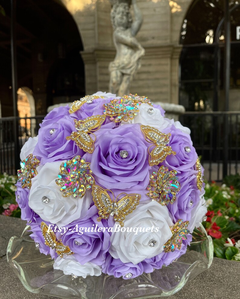 Pu&ograve; includere: Un bouquet da sposa fatto di rose bianche e lavanda con spille a forma di farfalla dorate e strass. Il bouquet &egrave; su un piatto di vetro trasparente. Il testo "Etsy-AguileraBouquets" &egrave; sul bouquet.