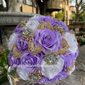 Pu&ograve; includere: Un bouquet da sposa con rose bianche e lavanda, adornato con spille a forma di farfalla dorate e strass iridescenti. Il bouquet &egrave; disposto in forma circolare e presenta una variet&agrave; di texture e colori.