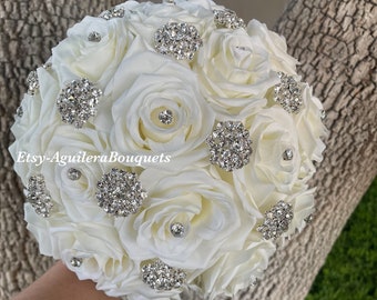 May include: A bridal bouquet featuring ivory roses adorned with sparkling rhinestone accents. The bouquet includes several large rhinestone brooches and smaller rhinestone embellishments, creating a glamorous and elegant look. The text "Etsy-Aguilera Bouquets" is visible.