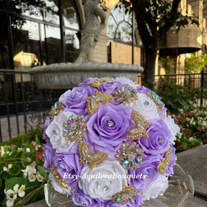 Pu&ograve; includere: Un bouquet di fiori di seta viola e bianco con farfalle dorate e strass. Il bouquet &egrave; su un piedistallo di vetro trasparente.