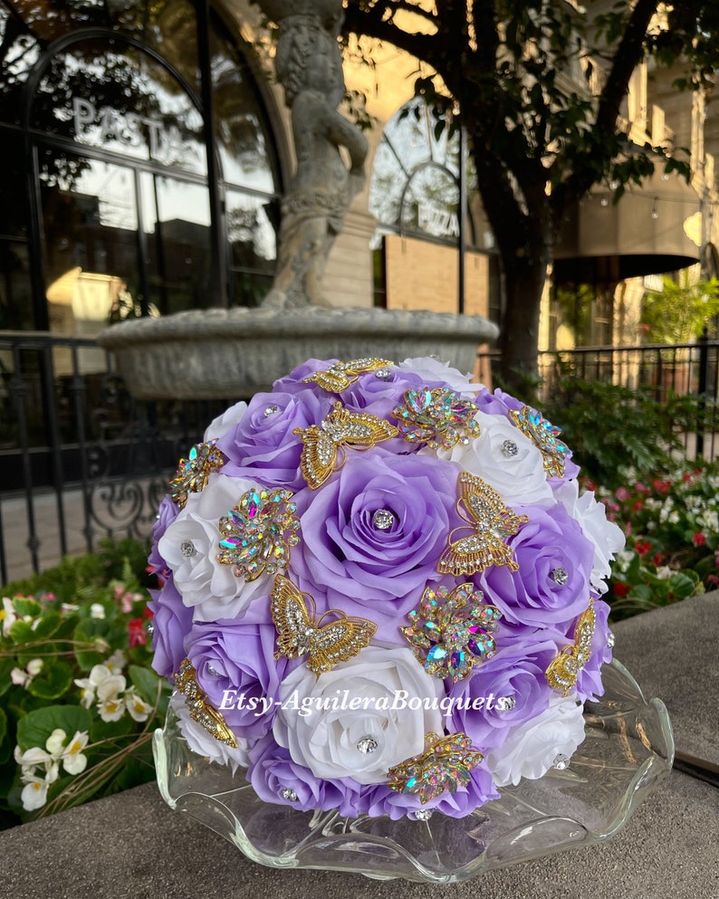 Pu&ograve; includere: Un bouquet di fiori di seta viola e bianco con farfalle dorate e strass. Il bouquet &egrave; su un piedistallo di vetro trasparente.