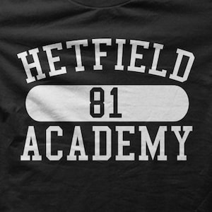May include: Black crew neck t-shirt with white text. The text reads "HETFIELD 81 ACADEMY" in a collegiate style font. The number "81" is centered within a white horizontal rectangle.