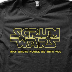 Könnte beinhalten: Schwarzes T-Shirt mit gelbem Text, der "SCRUM WARS" in einer stilisierten Schriftart und den Satz "MAY BRUTE FORCE BE WITH YOU" darunter zeigt. Das Shirt hat einen Rundhalsausschnitt und kurze Ärmel.