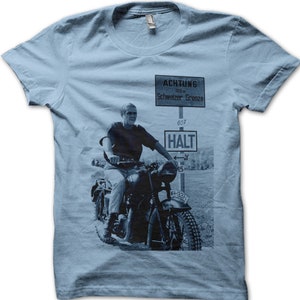 May include: A light blue t-shirt with a black and white image of a man riding a motorcycle. The image includes a sign that reads "ACHTUNG 100 m Schweizer Grenze 607 HALT".