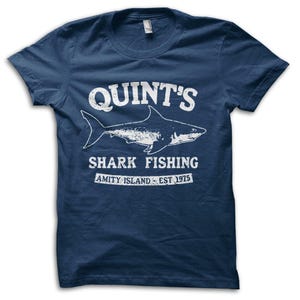 Könnte beinhalten: Marineblaues T-Shirt mit weißem Aufdruck. Der Aufdruck lautet "QUINT'S SHARK FISHING" mit einer Hai-Illustration und "AMITY ISLAND EST. 1975". Das T-Shirt hat einen Rundhalsausschnitt.