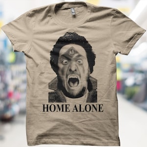 May include: A beige t-shirt with a black and white image of a man with a surprised expression and open mouth. The words "HOME ALONE" are printed in black below the image. The shirt has short sleeves and a round neck.