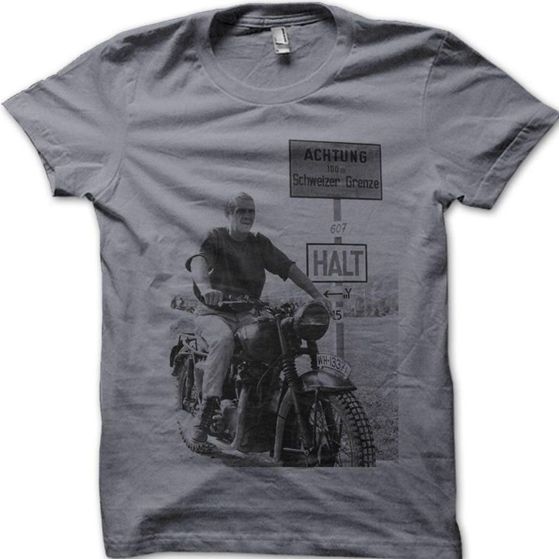 May include: A gray t-shirt with a black and white image of a man riding a motorcycle on a dirt road. The image includes a sign that reads "ACHTUNG 100 m Schweizer Grenze 607 HALT".