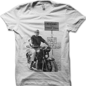 May include: A black and white photo of a man riding a motorcycle on a dirt road. The man is wearing a t-shirt and jeans. The motorcycle has a sidecar. There is a sign in the background that says "ACHTUNG 100 m Schweizer Grenze 607 HALT".