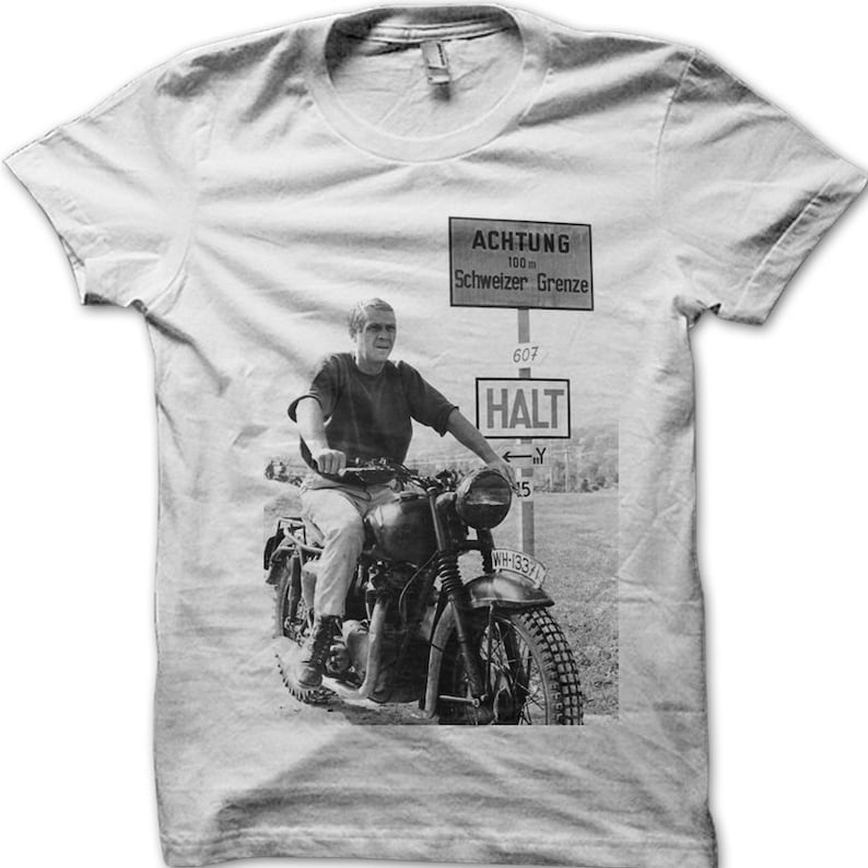 May include: A black and white photo of a man riding a motorcycle on a dirt road. The man is wearing a t-shirt and jeans. The motorcycle has a sidecar. There is a sign in the background that says "ACHTUNG 100 m Schweizer Grenze 607 HALT".