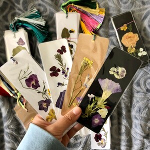 May include: Assortment of handmade bookmarks featuring pressed flowers on various colored backgrounds. The bookmarks are made with different types of flowers, including purple, yellow, and white.