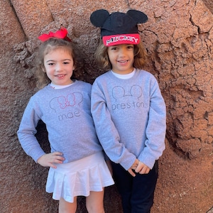 May include: Two children wearing gray sweatshirts with Mickey Mouse ear designs and names embroidered on them. One child wears a Minnie Mouse bow and a white skirt. The other child wears a Mickey Mouse hat with the word "Mickey" on it.