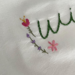 May include: Embroidered white fabric with the word "wild" in green thread. The word is surrounded by pink and purple flowers and green stems.