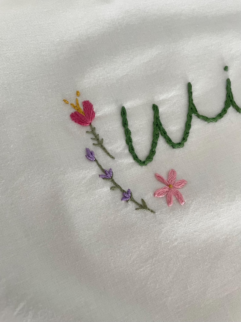 May include: Embroidered white fabric with the word "wild" in green thread. The word is surrounded by pink and purple flowers and green stems.
