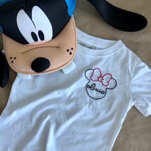 May include: White t-shirt with a black and red embroidered Minnie Mouse design and the name "Olivia" underneath.