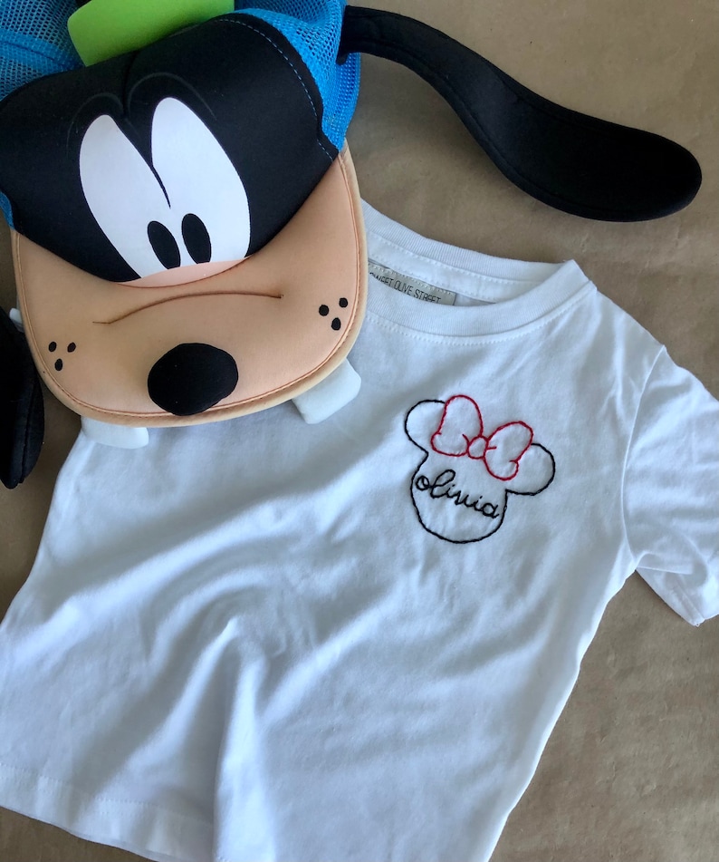 May include: White t-shirt with a black and red embroidered Minnie Mouse design and the name "Olivia" underneath.