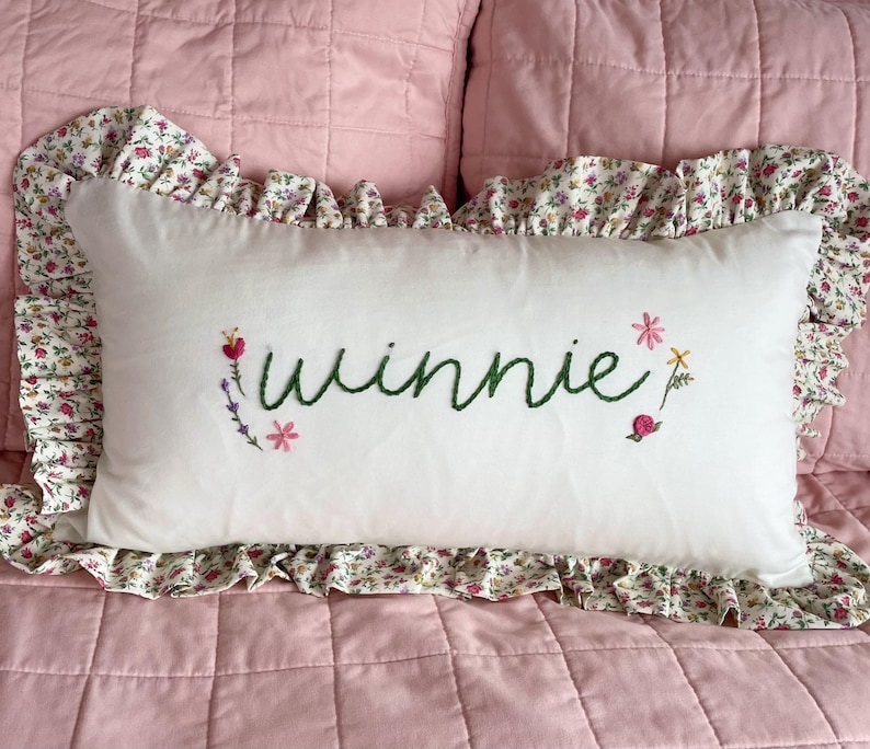 May include: A white pillow with a floral print border and the name "Winnie" embroidered in green thread. The pillow is decorated with small embroidered flowers in pink, purple, and green.