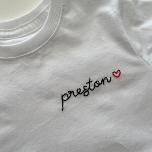 May include: White t-shirt with black embroidered text "preston" and a small red heart.