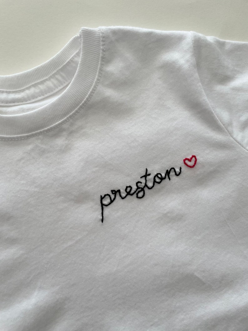 May include: White t-shirt with black embroidered text "preston" and a small red heart.