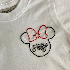May include: White t-shirt with a black and red embroidered Mickey Mouse head with the word "sissy" written inside.