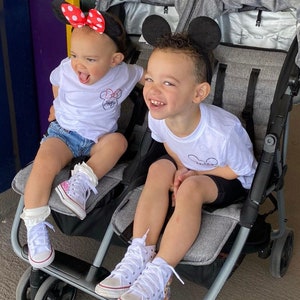 May include: Two children wearing Mickey Mouse ears sit in a double stroller. The child on the left is wearing a white shirt with a red and white Mickey Mouse design and denim shorts. The child on the right is wearing a white shirt with a black Mickey Mouse design and black shorts. Both children are wearing white high-top sneakers with colorful designs.