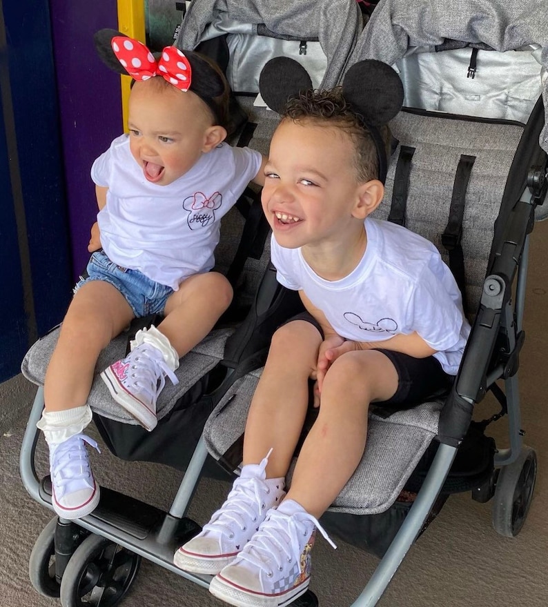 May include: Two children wearing Mickey Mouse ears sit in a double stroller. The child on the left is wearing a white shirt with a red and white Mickey Mouse design and denim shorts. The child on the right is wearing a white shirt with a black Mickey Mouse design and black shorts. Both children are wearing white high-top sneakers with colorful designs.